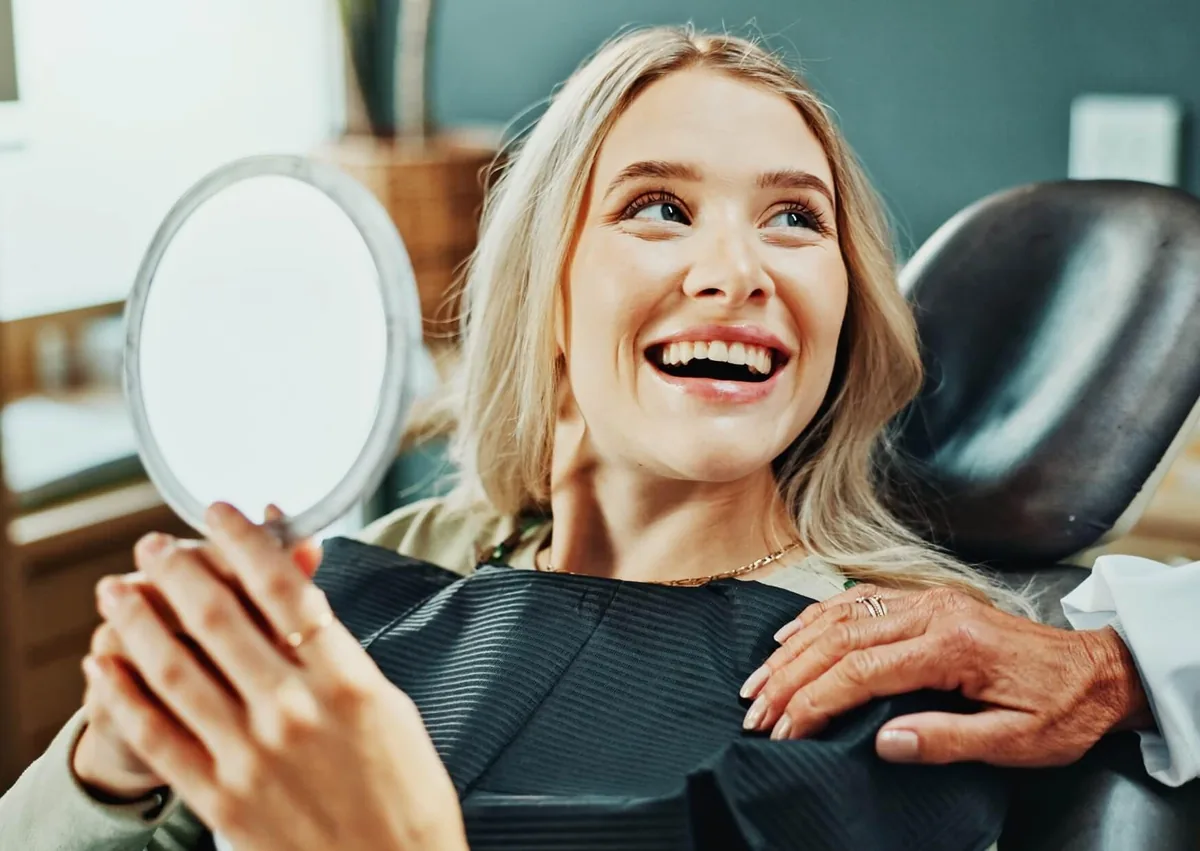 Smiling woman holding a mirror in a dental chair while admiring her smile makeover results after cosmetic dental treatment.